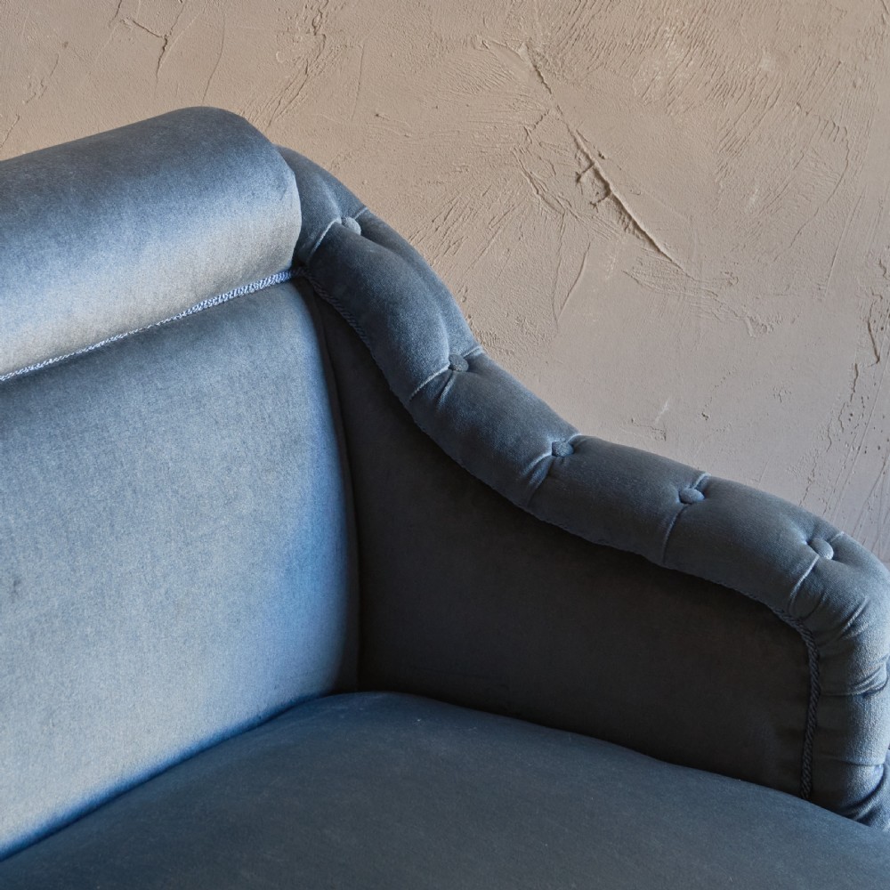 A Victorian bedroom settee upholstered in light blue velvet with buttoned details along the armrests and base, fitted with a tassel fringe skirt and turned wooden legs on castors. View X