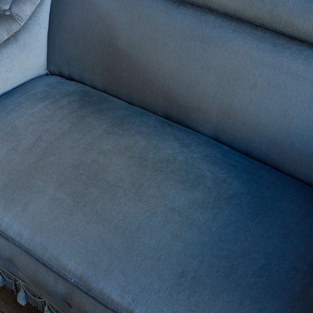 A Victorian bedroom settee upholstered in light blue velvet with buttoned details along the armrests and base, fitted with a tassel fringe skirt and turned wooden legs on castors. View XI