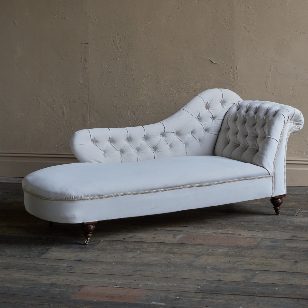 An antique Victorian chaise longue upholstered in off-white velvet with deep buttoning, a scrolled backrest, braided trim detailing, and dark turned legs on brass castors.