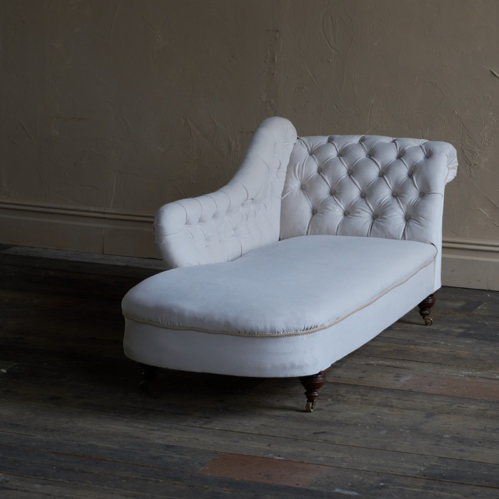 An antique Victorian chaise longue upholstered in off-white velvet with deep buttoning, a scrolled backrest, braided trim detailing, and dark turned legs on brass castors. View II
