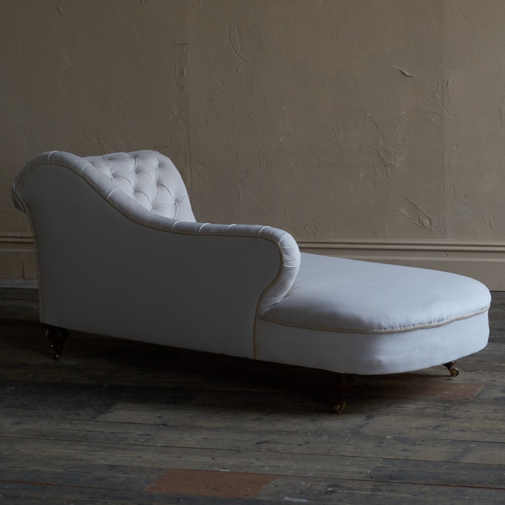 An antique Victorian chaise longue upholstered in off-white velvet with deep buttoning, a scrolled backrest, braided trim detailing, and dark turned legs on brass castors. View III