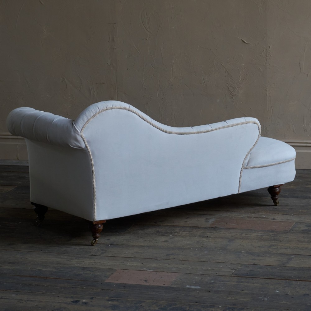 An antique Victorian chaise longue upholstered in off-white velvet with deep buttoning, a scrolled backrest, braided trim detailing, and dark turned legs on brass castors. View V