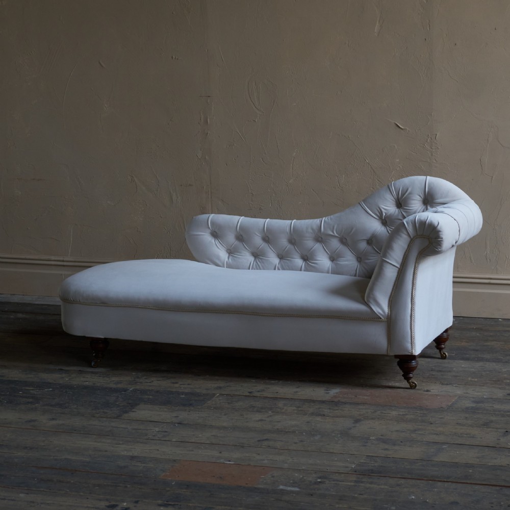 An antique Victorian chaise longue upholstered in off-white velvet with deep buttoning, a scrolled backrest, braided trim detailing, and dark turned legs on brass castors. View VI