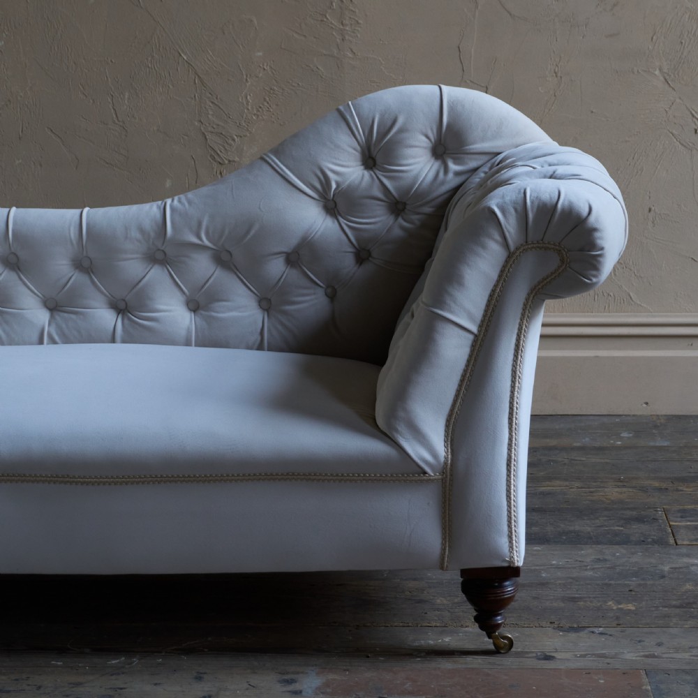 An antique Victorian chaise longue upholstered in off-white velvet with deep buttoning, a scrolled backrest, braided trim detailing, and dark turned legs on brass castors. View VII
