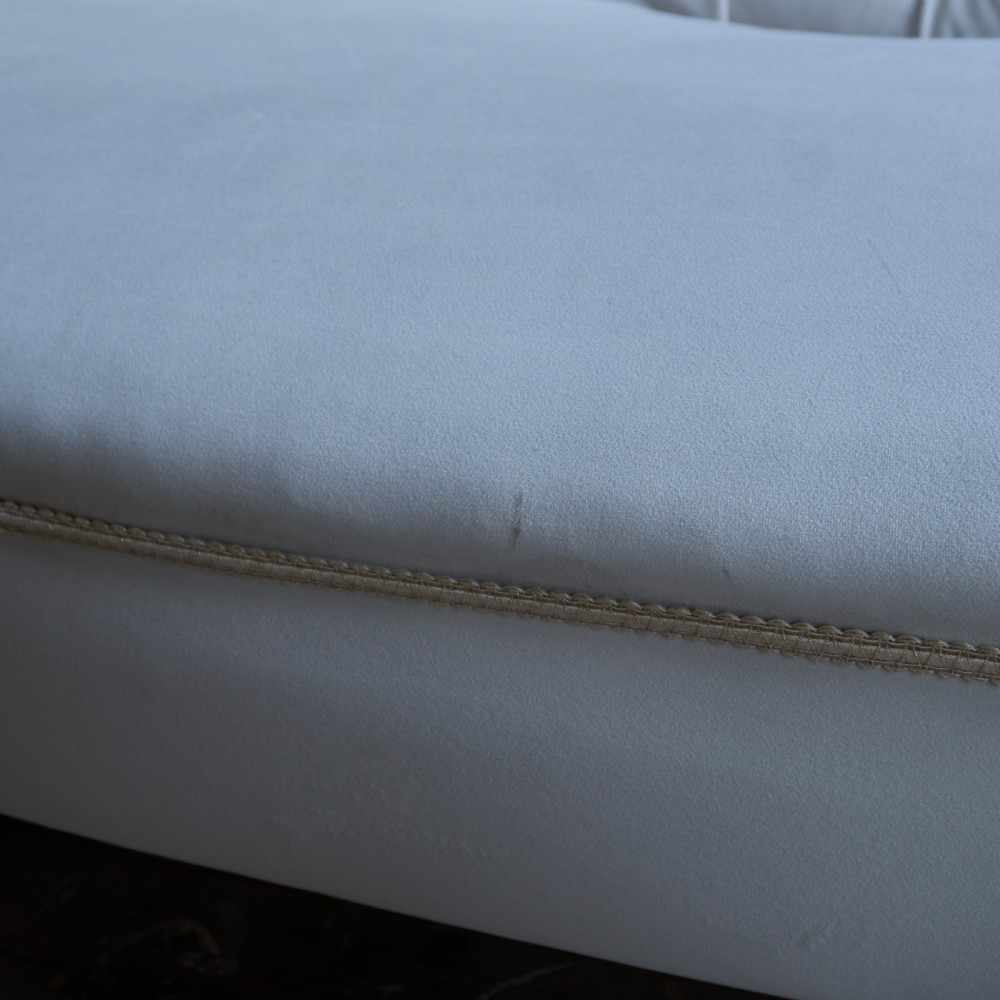An antique Victorian chaise longue upholstered in off-white velvet with deep buttoning, a scrolled backrest, braided trim detailing, and dark turned legs on brass castors. View VIII