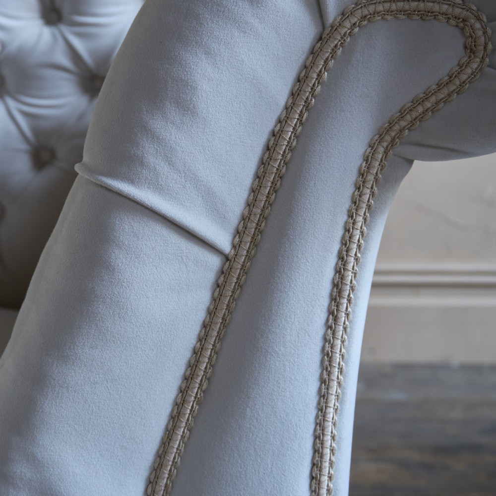 An antique Victorian chaise longue upholstered in off-white velvet with deep buttoning, a scrolled backrest, braided trim detailing, and dark turned legs on brass castors. View IX