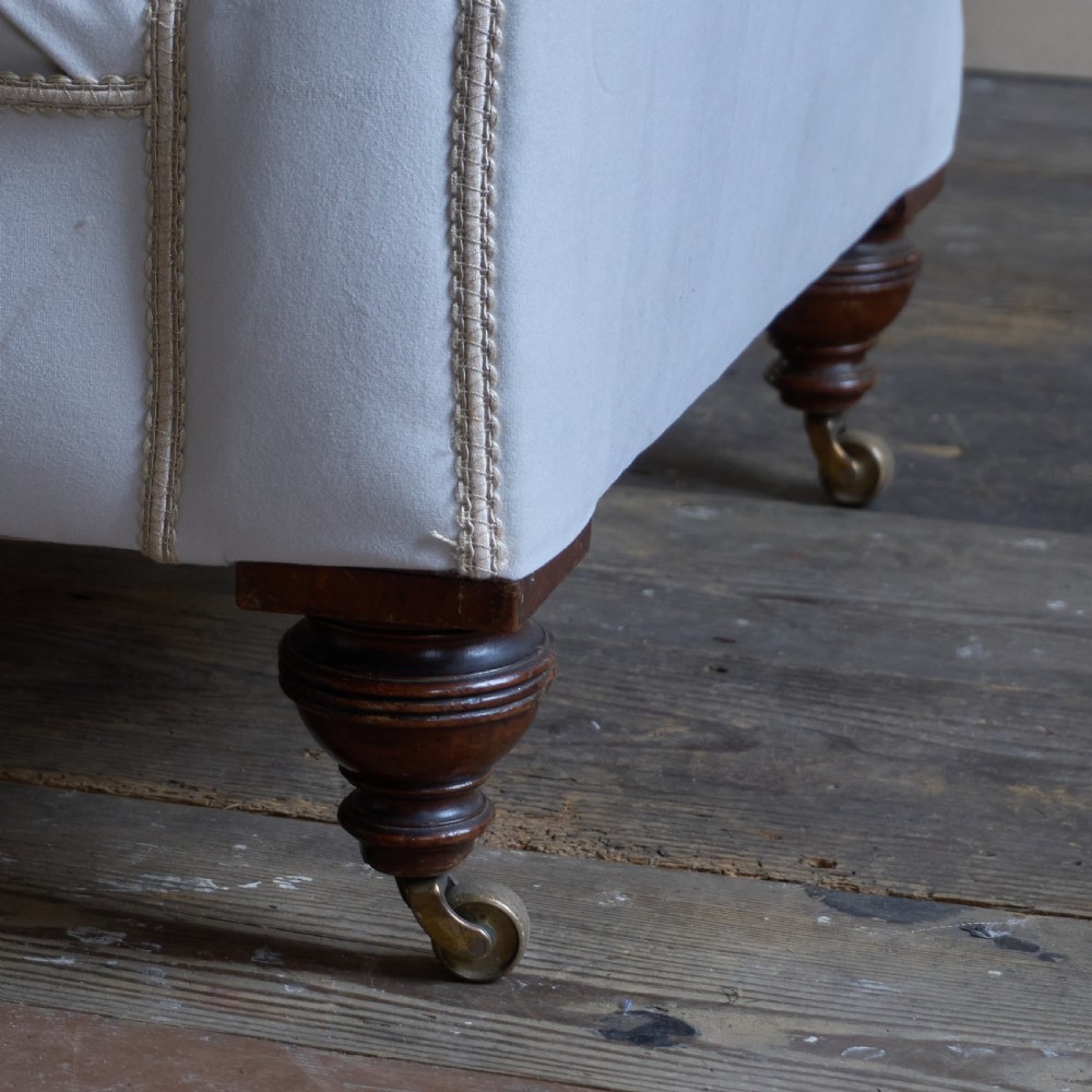 An antique Victorian chaise longue upholstered in off-white velvet with deep buttoning, a scrolled backrest, braided trim detailing, and dark turned legs on brass castors. View X