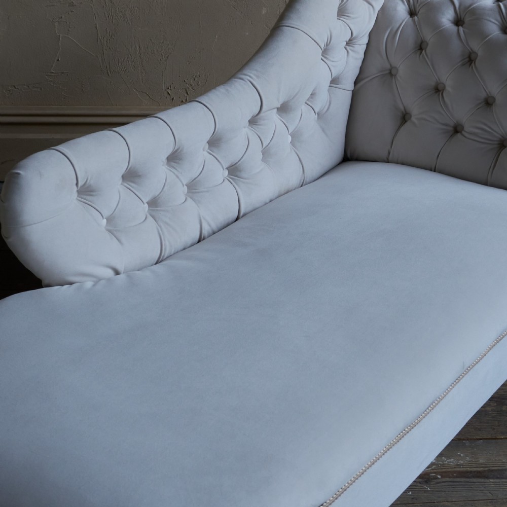 An antique Victorian chaise longue upholstered in off-white velvet with deep buttoning, a scrolled backrest, braided trim detailing, and dark turned legs on brass castors. View XI