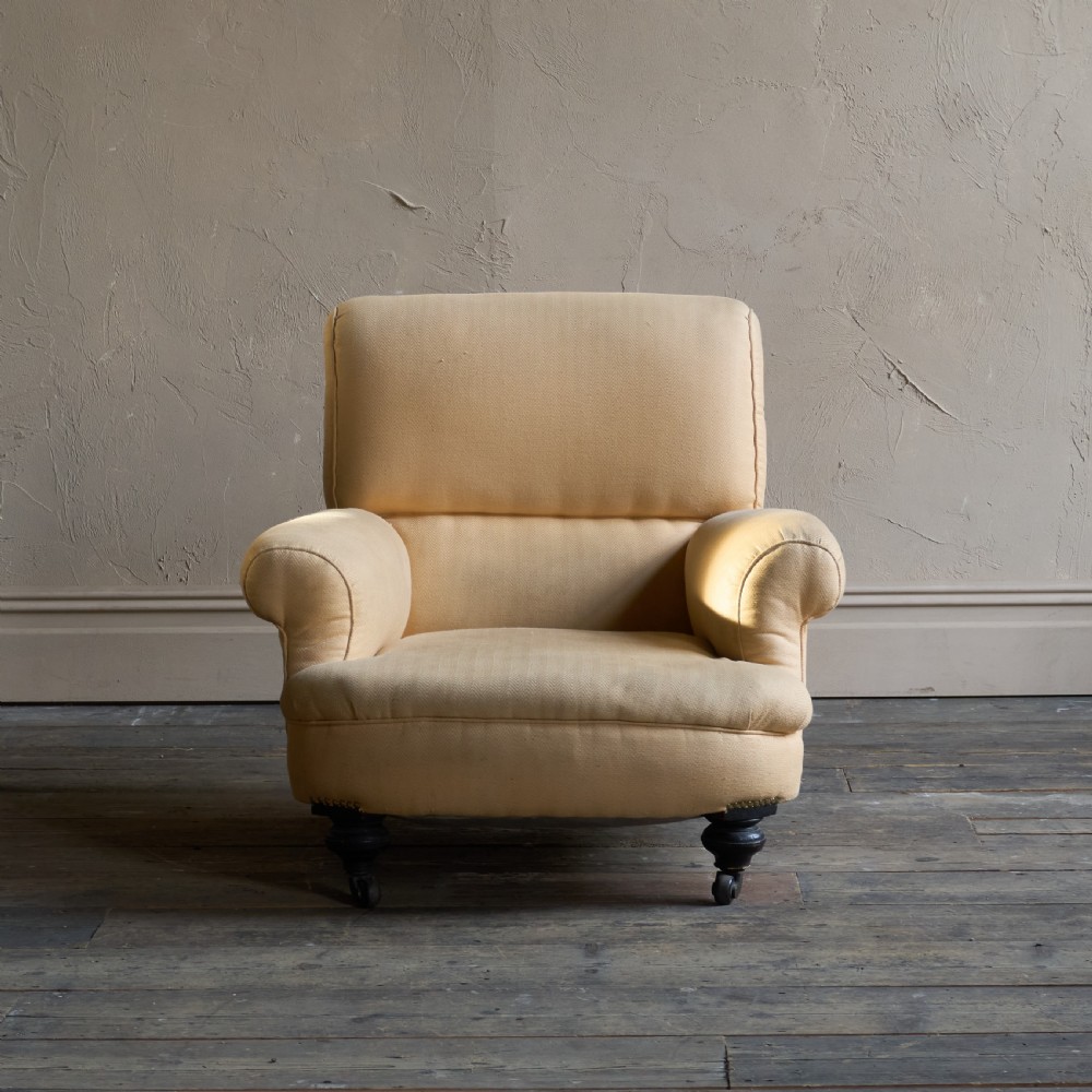 A deep-seated antique armchair with buff yellow fabric upholstery, scrolled arms, and dark turned wooden legs on castors. View III