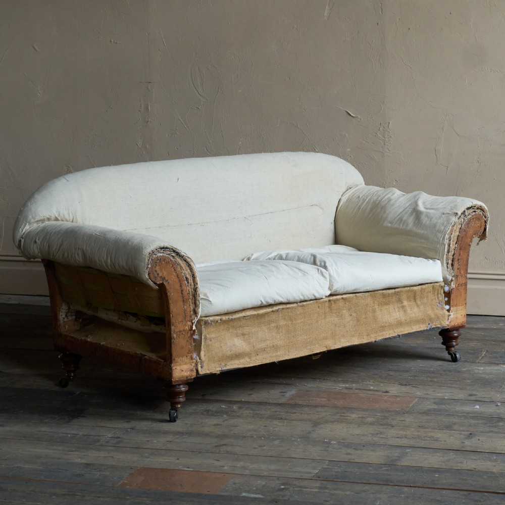 An antique two-seater country house sofa upholstered in unbleached calico with scroll arms and exposed wooden caster legs.