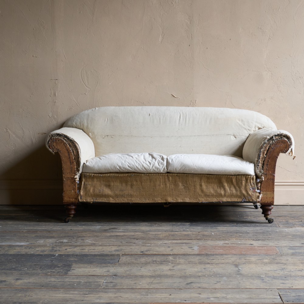 An antique two-seater country house sofa upholstered in unbleached calico with scroll arms and exposed wooden caster legs. View III