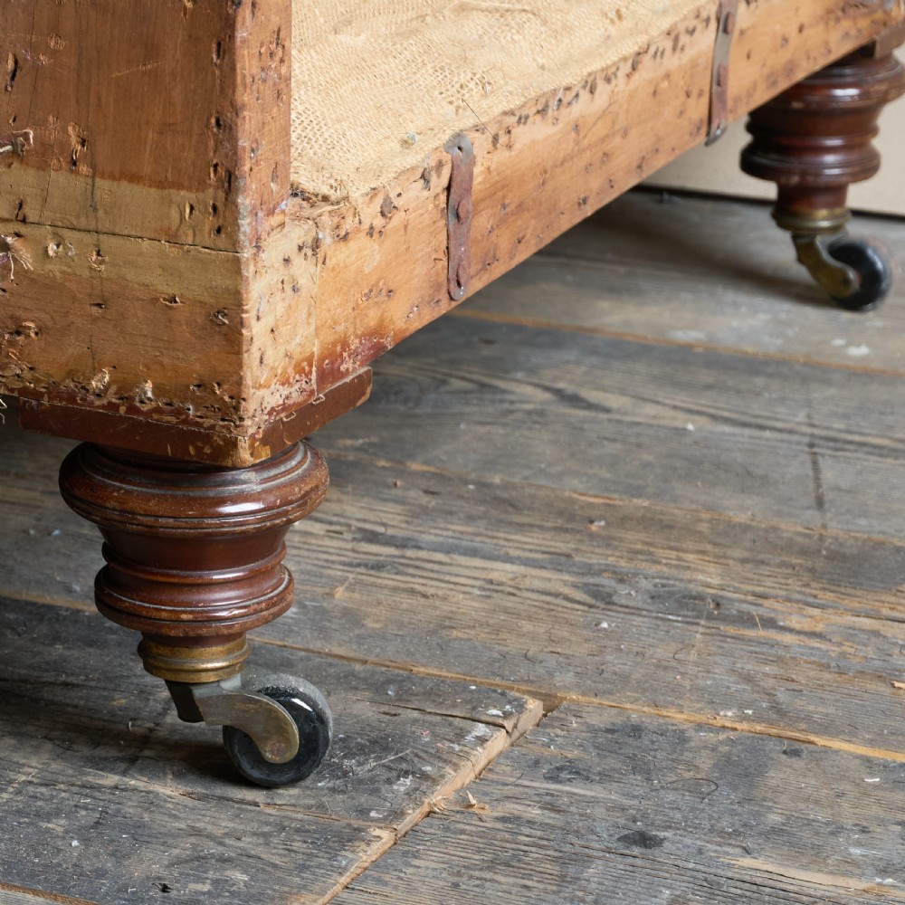 An antique two-seater country house sofa upholstered in unbleached calico with scroll arms and exposed wooden caster legs. View V