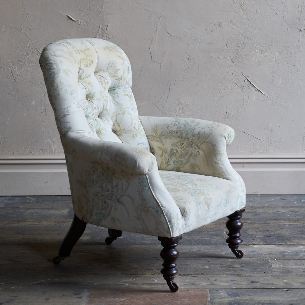 A Victorian spoon back armchair upholstered in light cream fabric with subtle floral patterns, featuring deep button tufting and turned dark wood legs on castors.