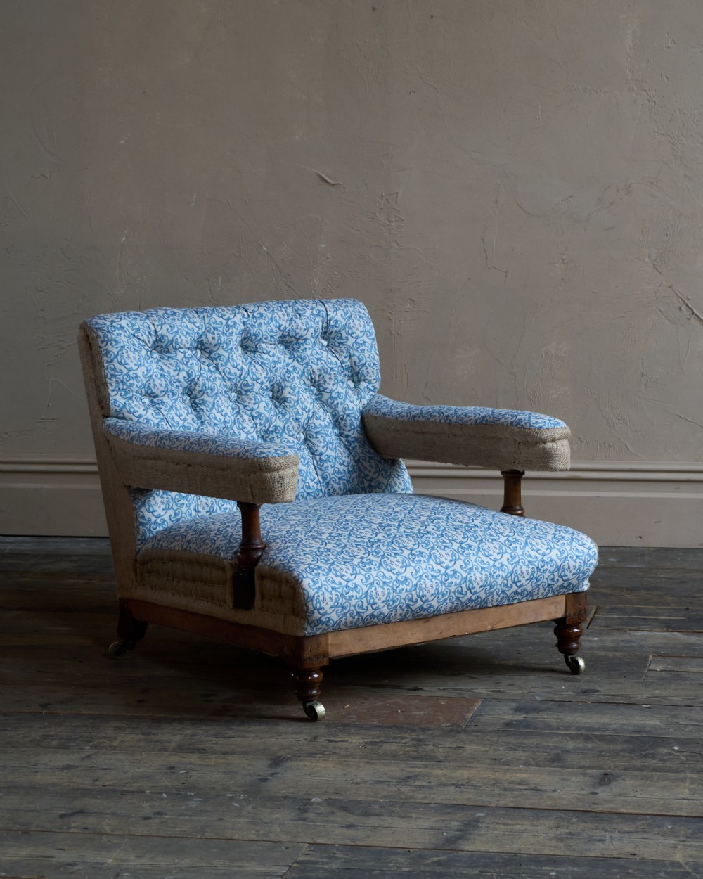 A Victorian open armchair upholstered in blue and white printed fabric with a button-tufted backrest, exposed Hessian sides, turned timber legs, and brass castors.