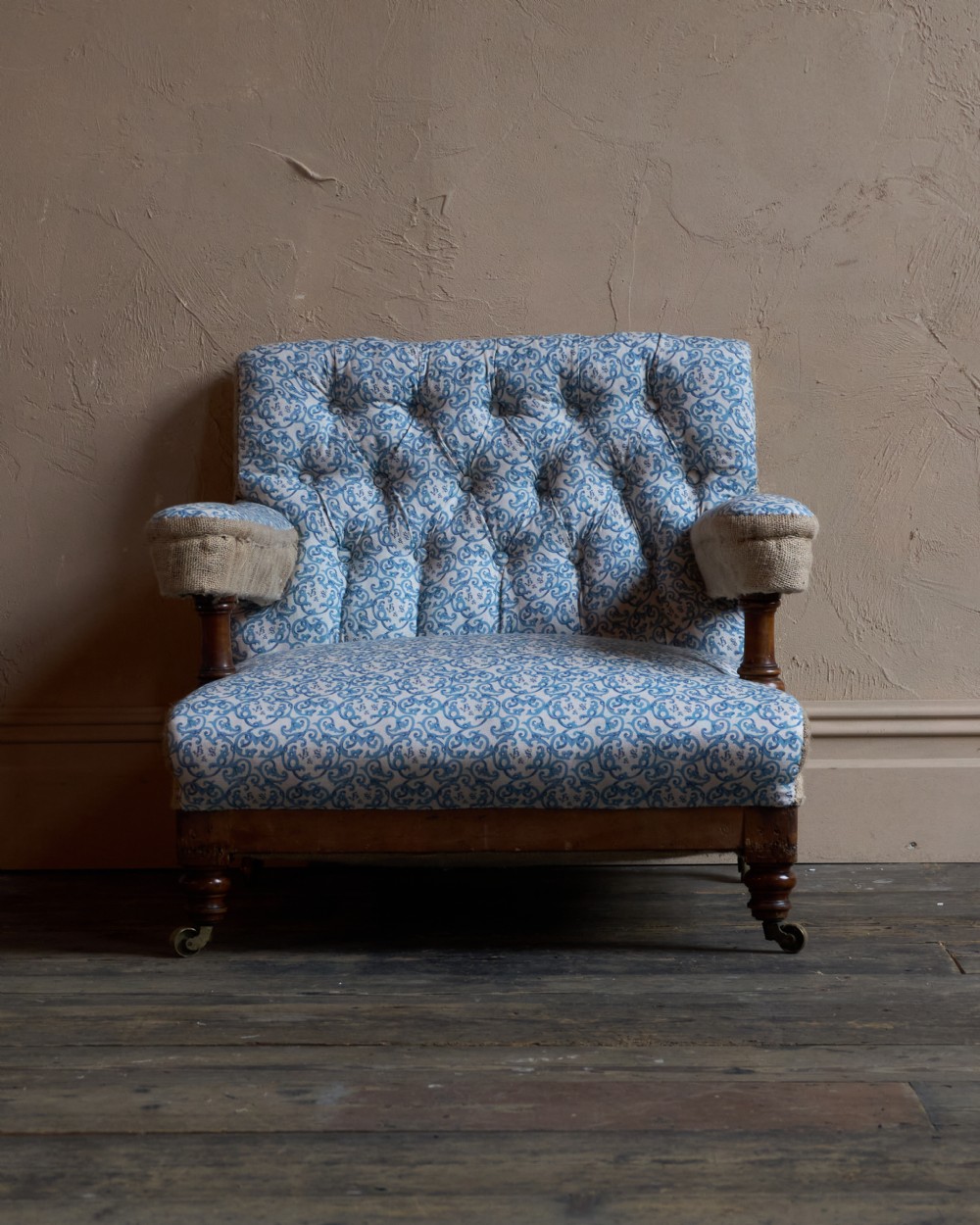 A Victorian open armchair upholstered in blue and white printed fabric with a button-tufted backrest, exposed Hessian sides, turned timber legs, and brass castors. View III