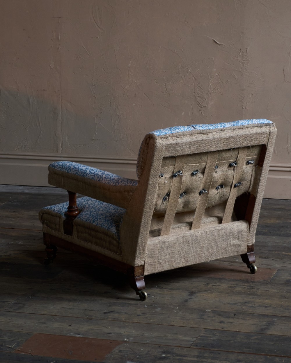 A Victorian open armchair upholstered in blue and white printed fabric with a button-tufted backrest, exposed Hessian sides, turned timber legs, and brass castors. View V