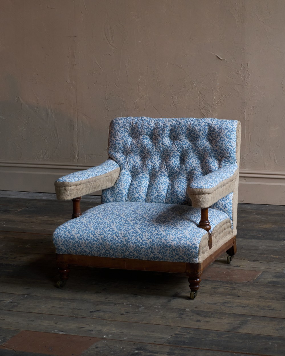 A Victorian open armchair upholstered in blue and white printed fabric with a button-tufted backrest, exposed Hessian sides, turned timber legs, and brass castors. View VI