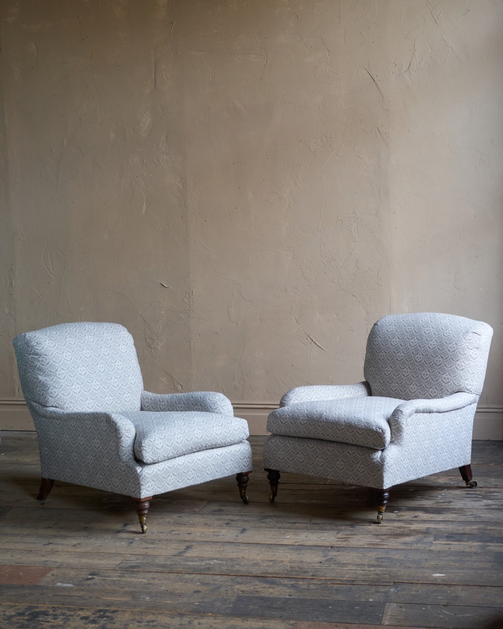 A pair of Victorian Howard and Sons Grafton style armchairs upholstered in patterned grey fabric with turned timber legs on brass castors.