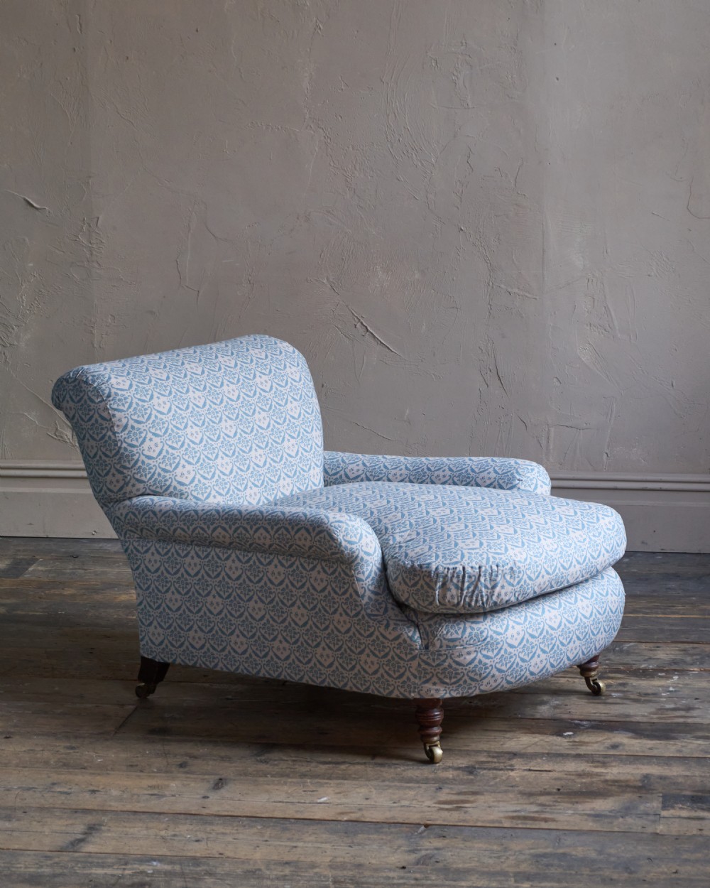 A low-slung, deeply cushioned club armchair upholstered in light blue and white patterned fabric with turned wooden legs on brass castors.
