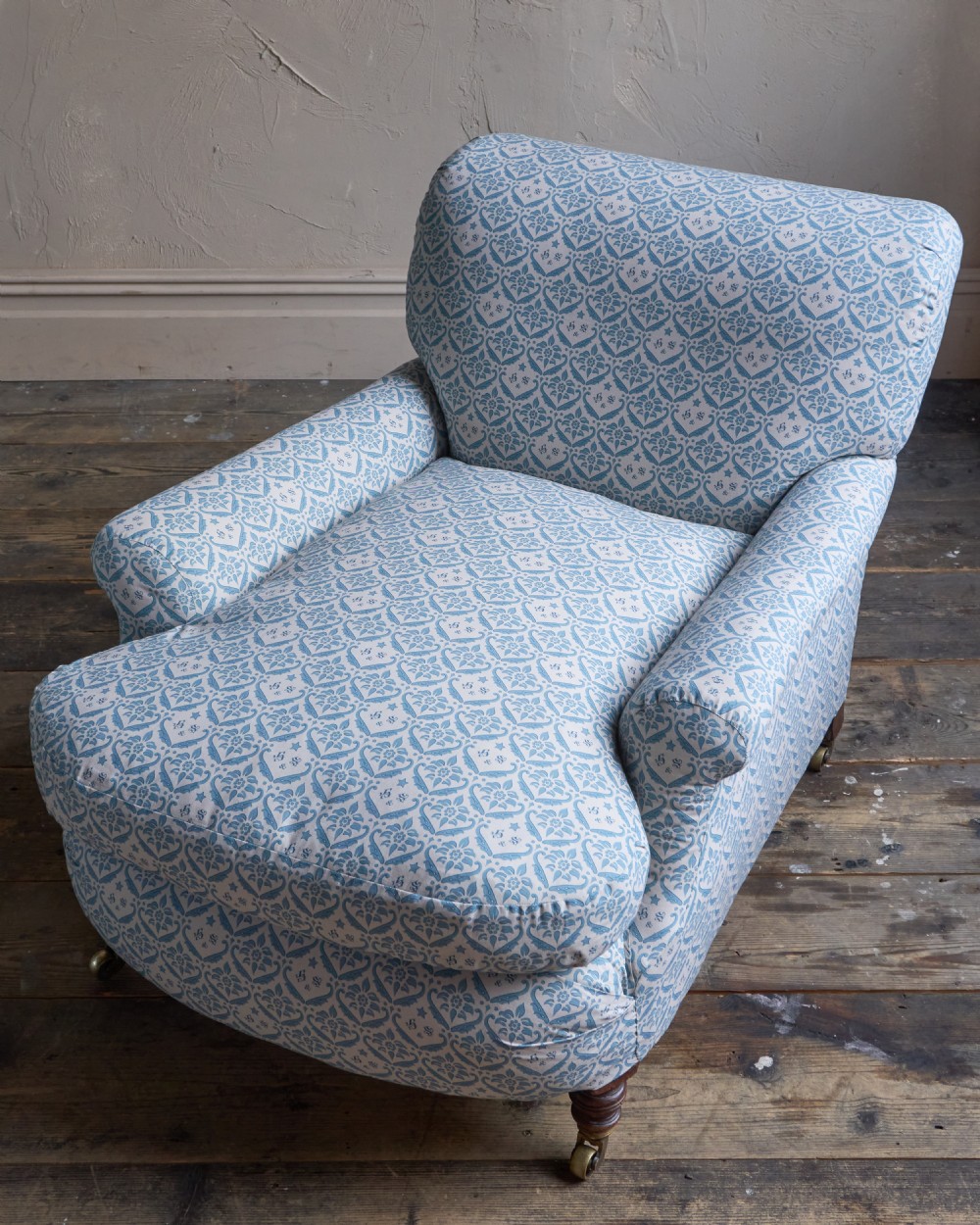 A low-slung, deeply cushioned club armchair upholstered in light blue and white patterned fabric with turned wooden legs on brass castors. View V
