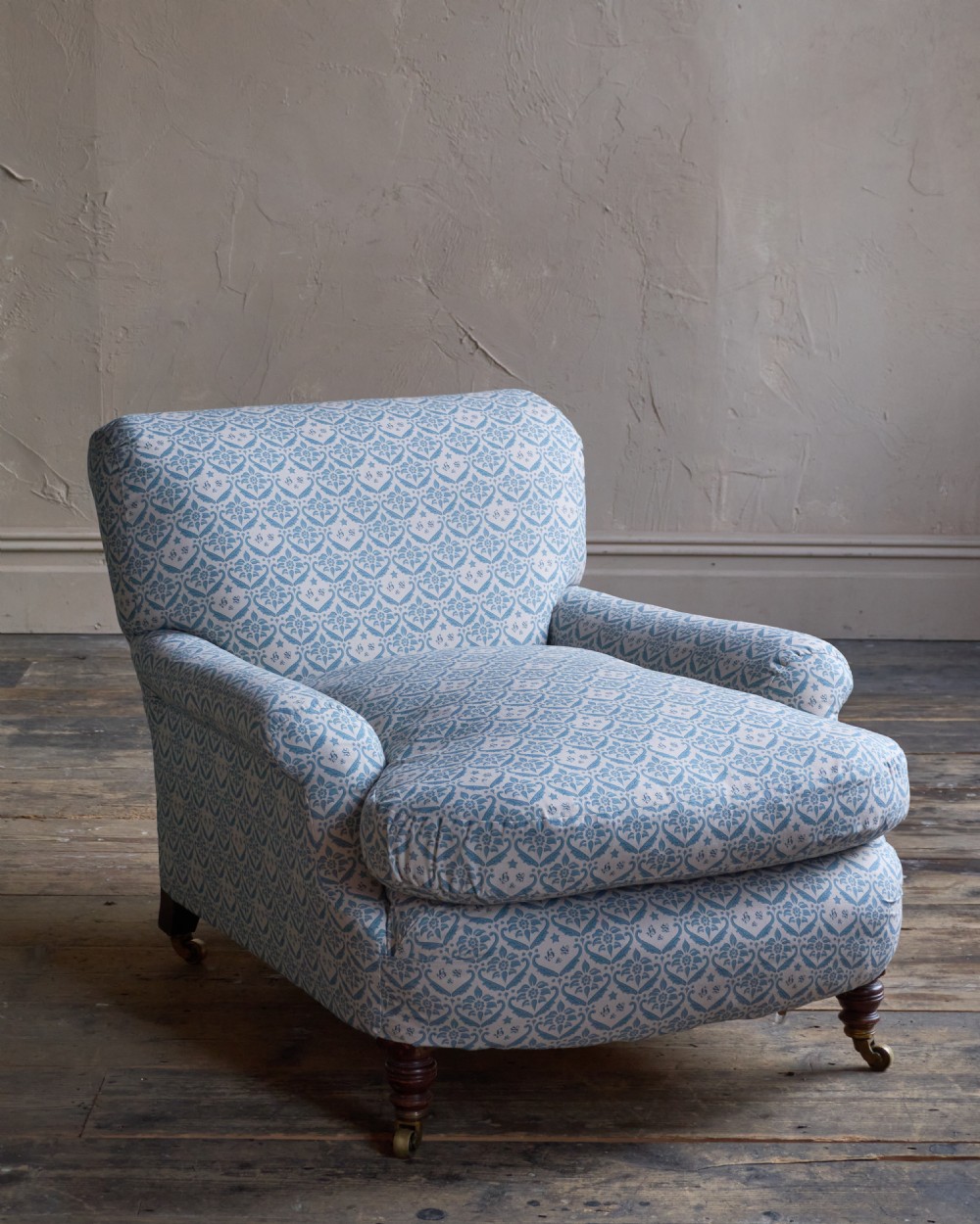 A low-slung, deeply cushioned club armchair upholstered in light blue and white patterned fabric with turned wooden legs on brass castors. View VI