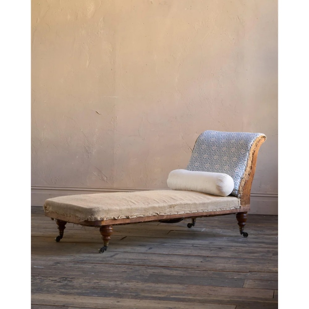 A 19th-century upholstered chaise longue with turned wooden legs on brass castors, patterned fabric backrest, hessian base, and cream bolster cushion. View III