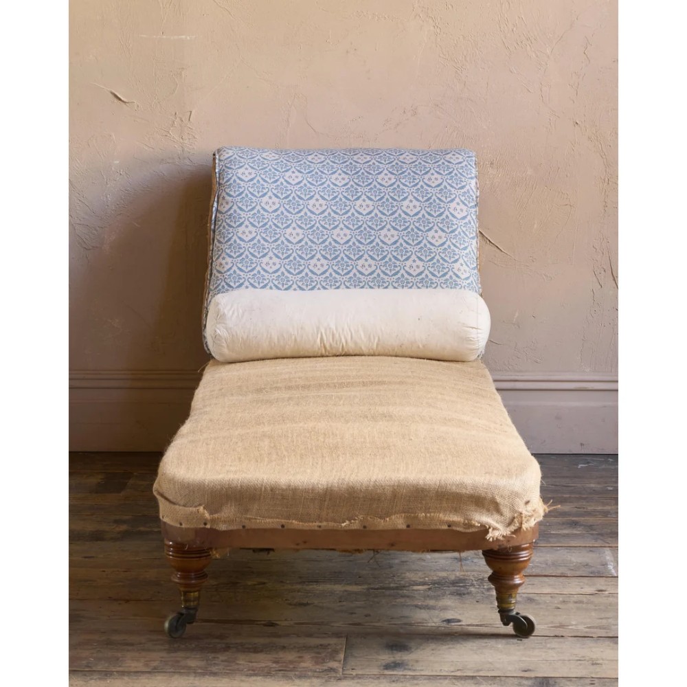 A 19th-century upholstered chaise longue with turned wooden legs on brass castors, patterned fabric backrest, hessian base, and cream bolster cushion. View V