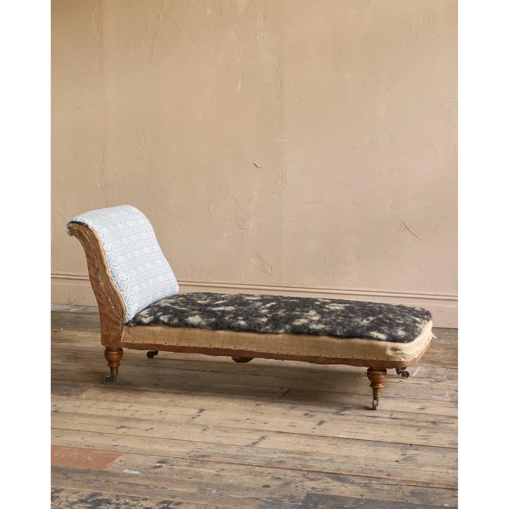 A 19th-century upholstered chaise longue with turned wooden legs on brass castors, patterned fabric backrest, hessian base, and cream bolster cushion. View X