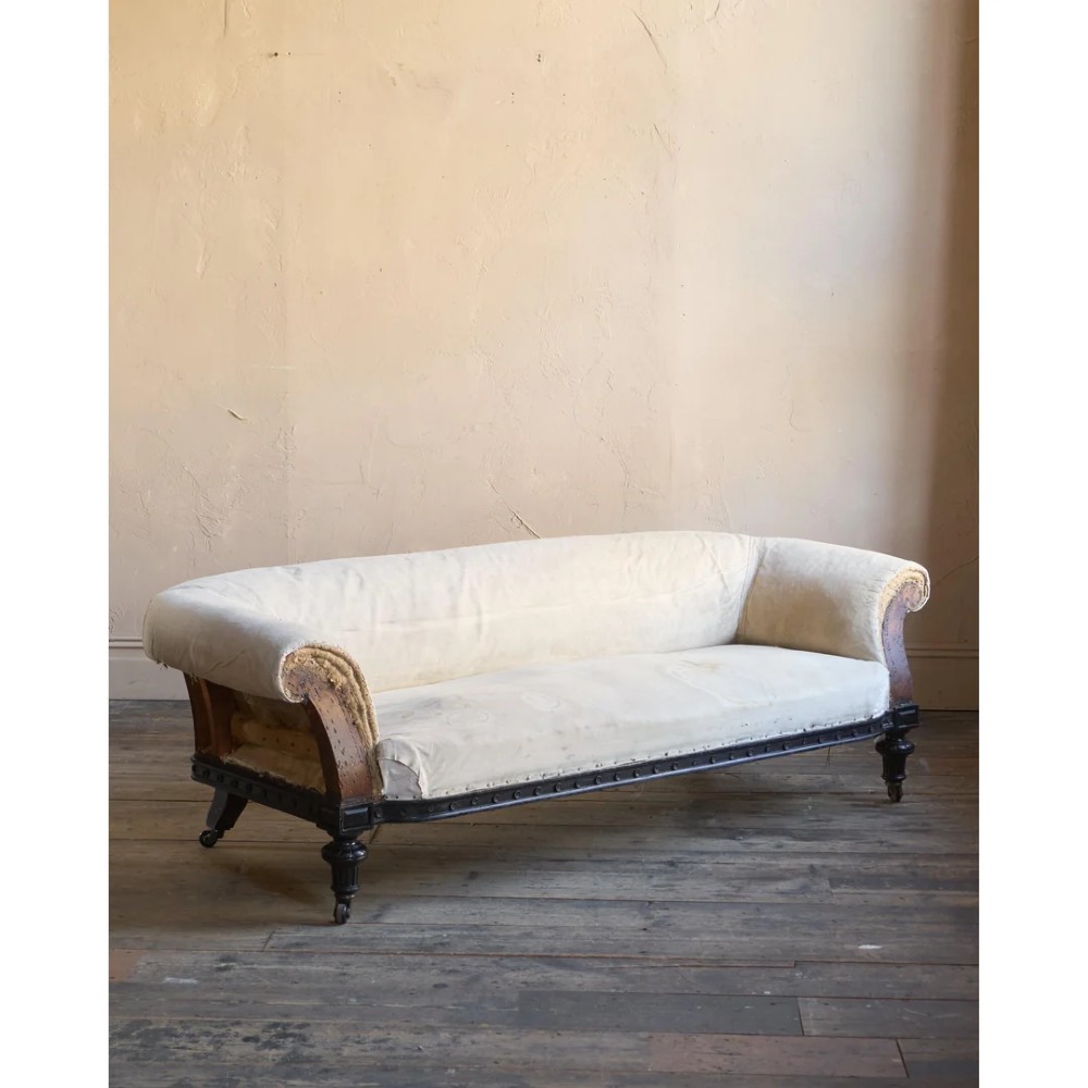 An antique Aesthetic Movement Chesterfield sofa featuring off-white upholstery, rolled arms, a dark carved timber frame, and turned legs with castors.