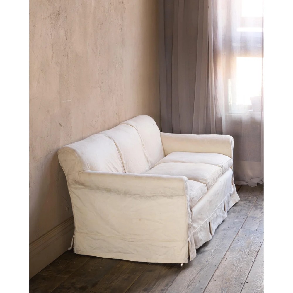 An Edwardian upholstered sofa featuring off-white neutral fabric, low padded armrests, deep seat cushions, and a pleated base skirt.