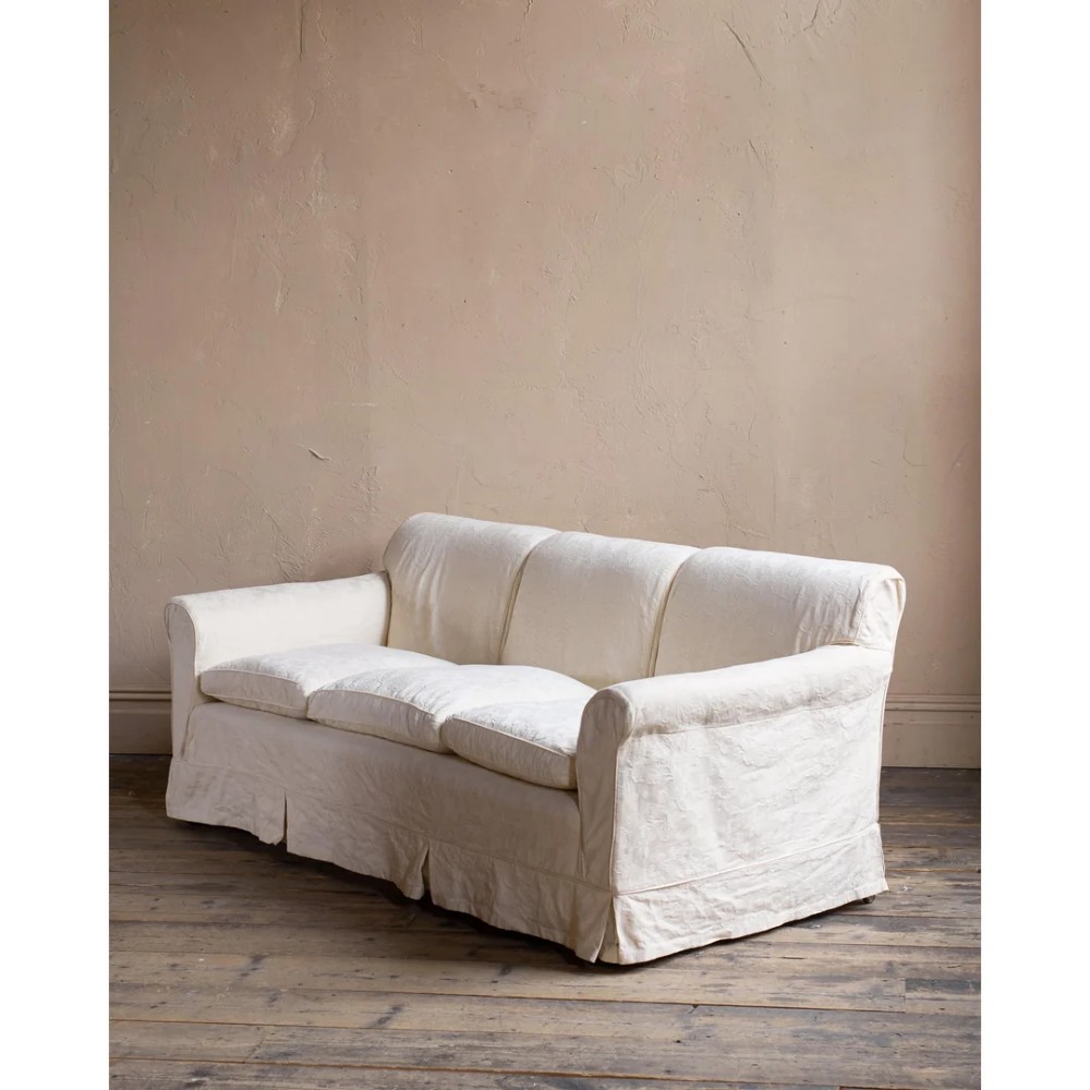 An Edwardian upholstered sofa featuring off-white neutral fabric, low padded armrests, deep seat cushions, and a pleated base skirt. View VII
