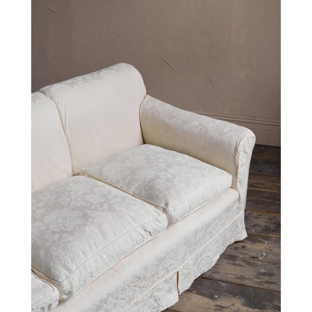 An Edwardian upholstered sofa featuring off-white neutral fabric, low padded armrests, deep seat cushions, and a pleated base skirt. View IX