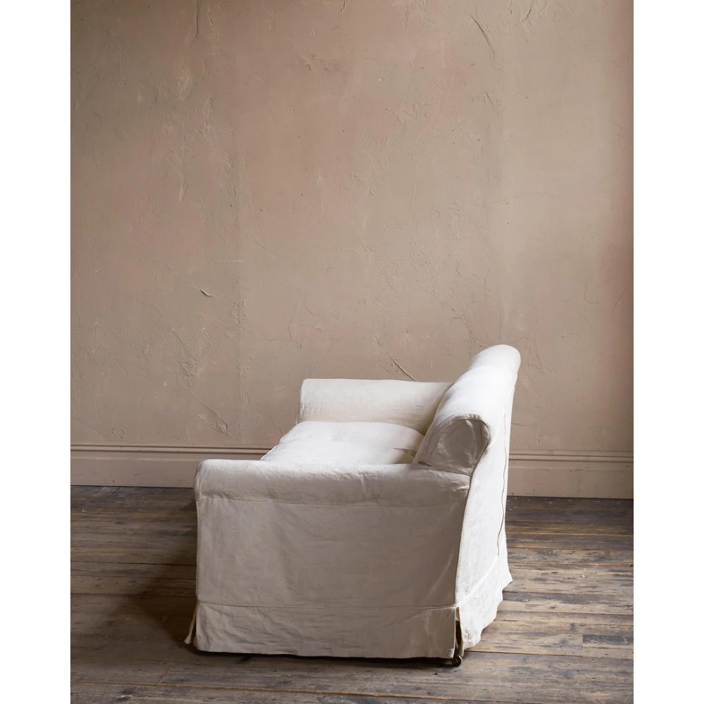 An Edwardian upholstered sofa featuring off-white neutral fabric, low padded armrests, deep seat cushions, and a pleated base skirt. View X