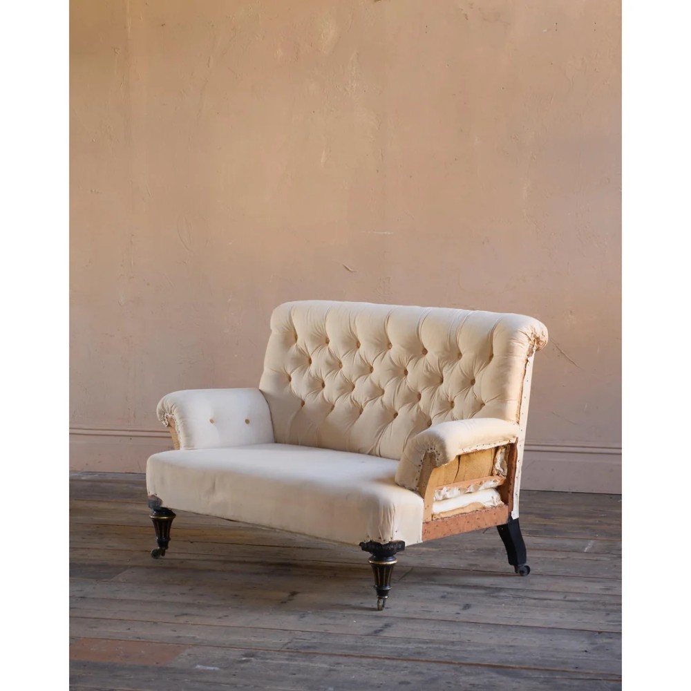 A Victorian two-seater sofa featuring deep button-tufted off-white calico upholstery, scroll arms, and turned front legs with brass castors.