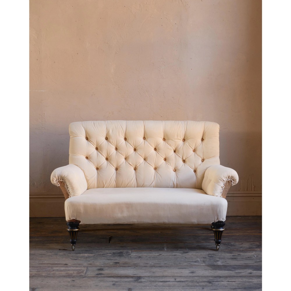 A Victorian two-seater sofa featuring deep button-tufted off-white calico upholstery, scroll arms, and turned front legs with brass castors. View V