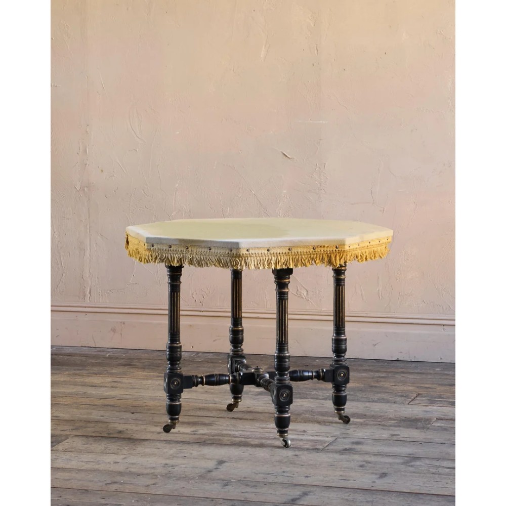 An octagonal Victorian centre table with ebonised turned legs, brass castors, and a pale top trimmed with fringe.
