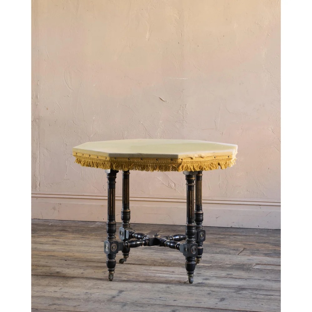 An octagonal Victorian centre table with ebonised turned legs, brass castors, and a pale top trimmed with fringe. View II