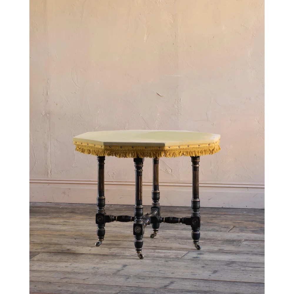 An octagonal Victorian centre table with ebonised turned legs, brass castors, and a pale top trimmed with fringe. View III