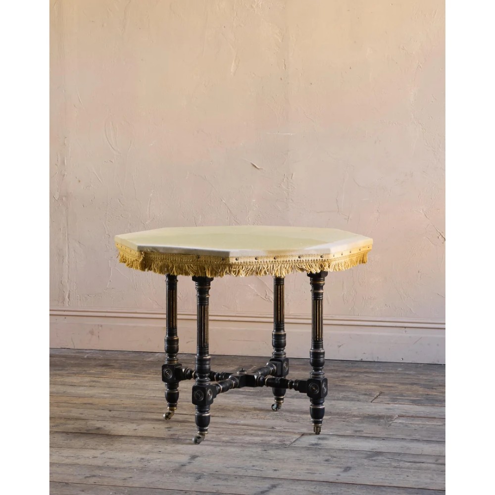An octagonal Victorian centre table with ebonised turned legs, brass castors, and a pale top trimmed with fringe. View IV