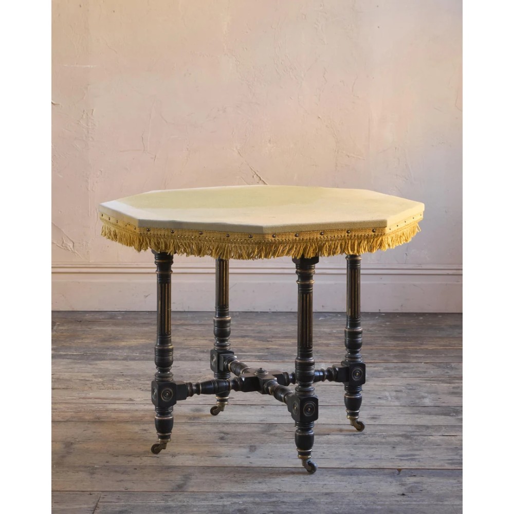 An octagonal Victorian centre table with ebonised turned legs, brass castors, and a pale top trimmed with fringe. View V