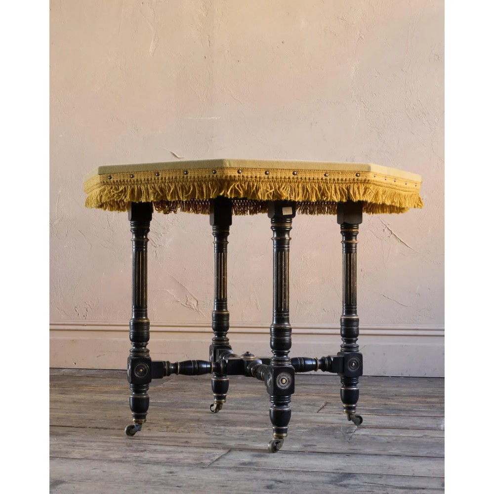 An octagonal Victorian centre table with ebonised turned legs, brass castors, and a pale top trimmed with fringe. View VI