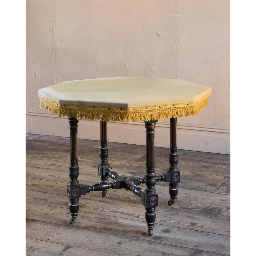 An octagonal Victorian centre table with ebonised turned legs, brass castors, and a pale top trimmed with fringe. View VII