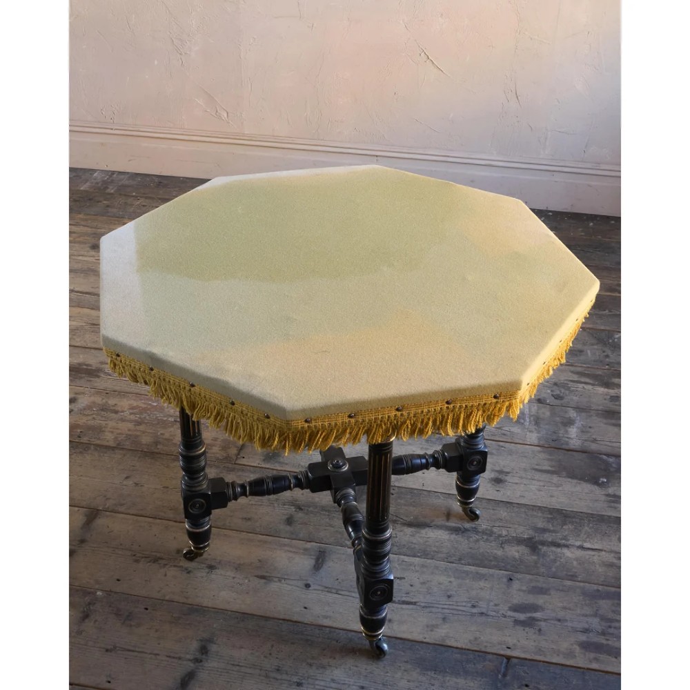 An octagonal Victorian centre table with ebonised turned legs, brass castors, and a pale top trimmed with fringe. View VIII