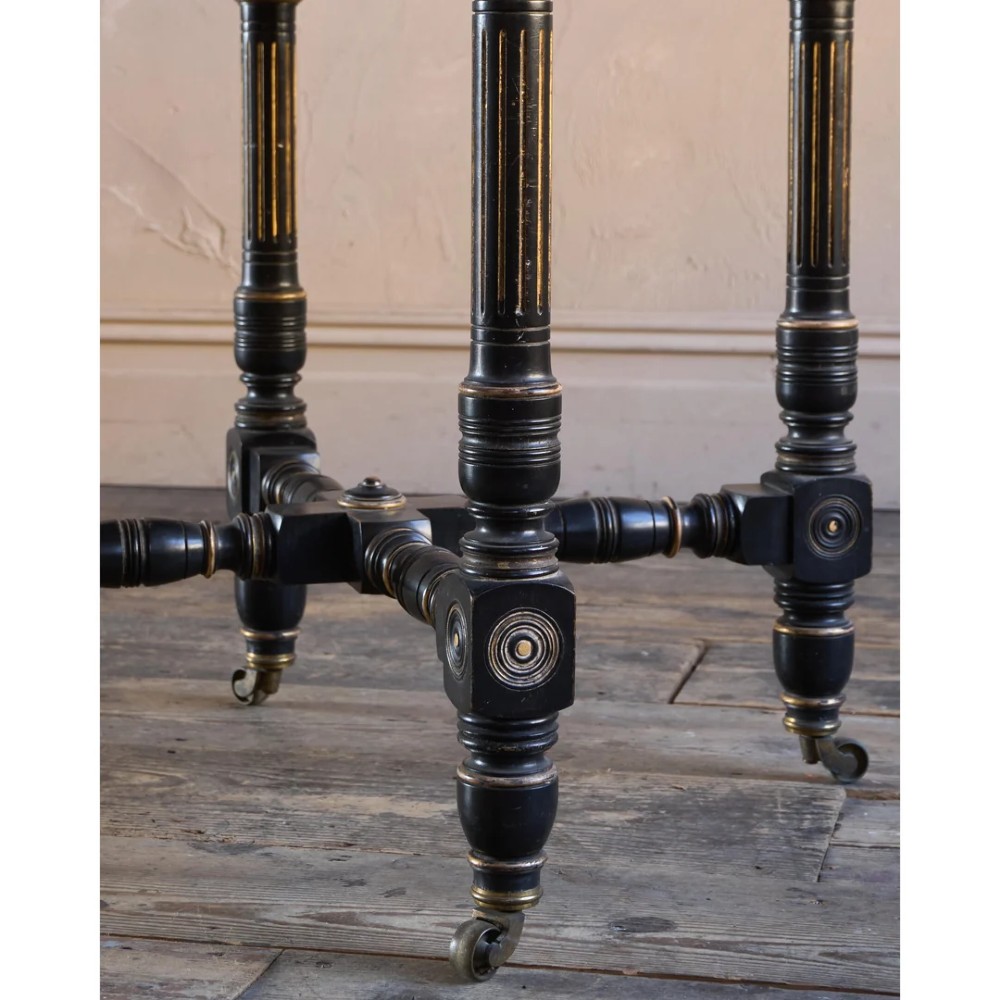 An octagonal Victorian centre table with ebonised turned legs, brass castors, and a pale top trimmed with fringe. View IX