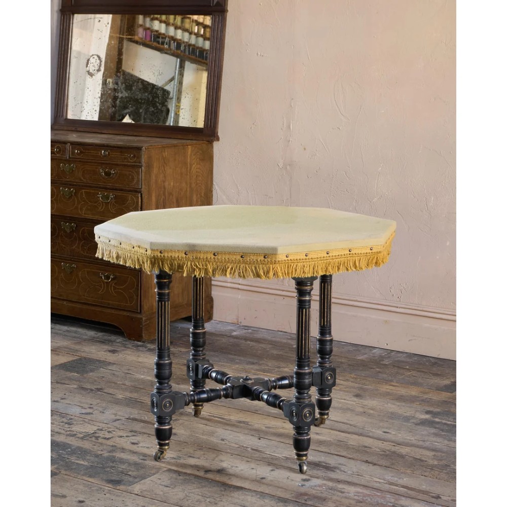 An octagonal Victorian centre table with ebonised turned legs, brass castors, and a pale top trimmed with fringe. View X