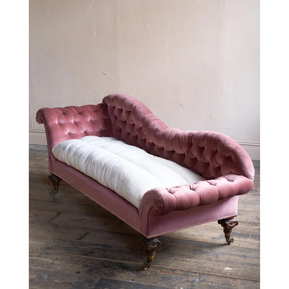 A Victorian deep-buttoned rose pink velvet chaise longue featuring carved mahogany Ionic column legs on brass castors with a loose white seat cushion.