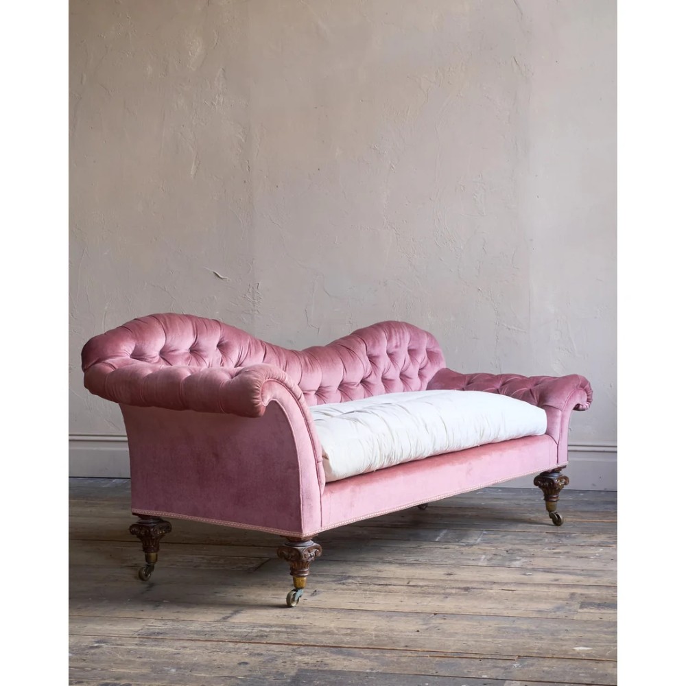 A Victorian deep-buttoned rose pink velvet chaise longue featuring carved mahogany Ionic column legs on brass castors with a loose white seat cushion. View II
