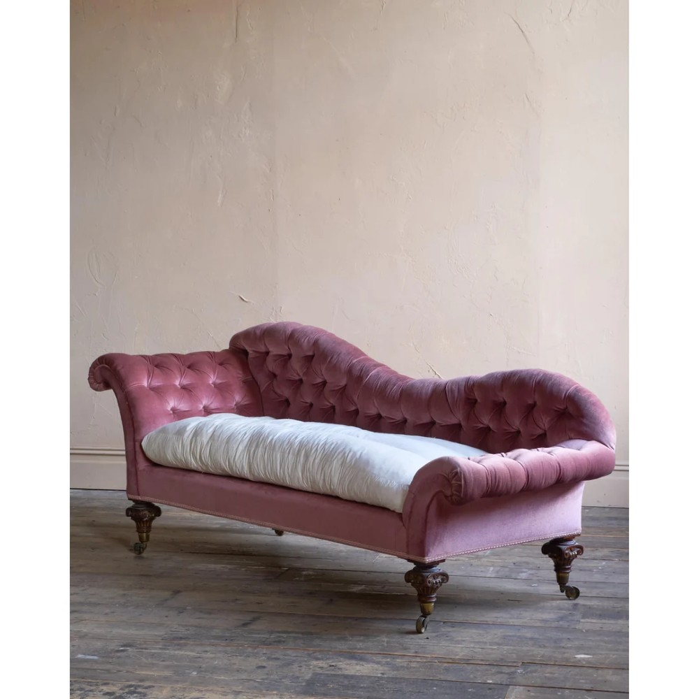 A Victorian deep-buttoned rose pink velvet chaise longue featuring carved mahogany Ionic column legs on brass castors with a loose white seat cushion. View V