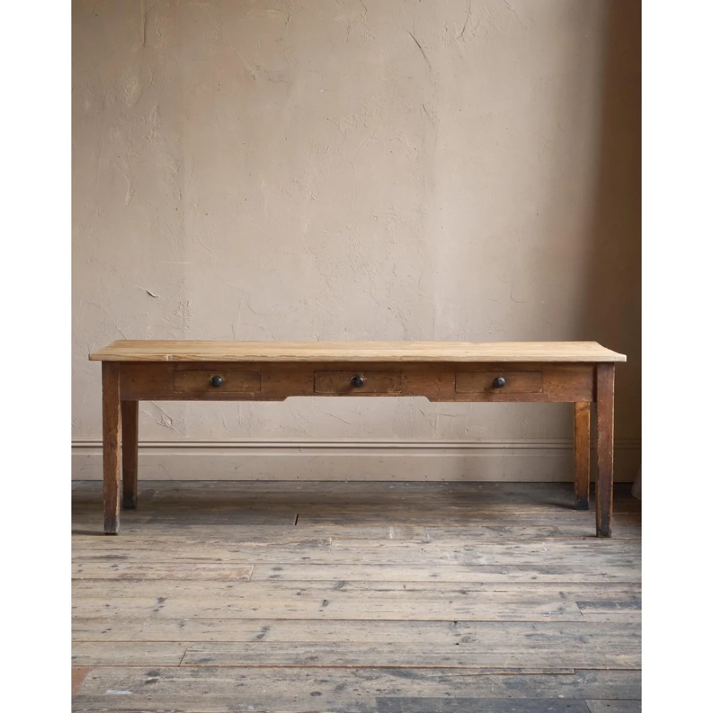 A long natural pine scullery table featuring three drawer fronts with dark wooden knobs and straight legs.