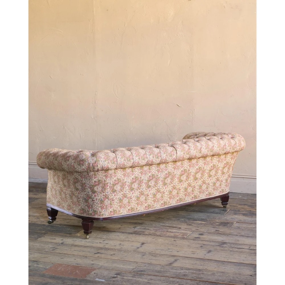 A nineteenth-century Chesterfield sofa with patterned floral upholstery, deep button tufting on the backrest and rolled arms, loose seat cushions, and turned legs on brass castors. View V